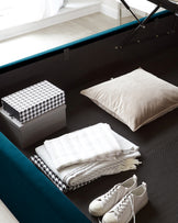 A modern, multifunctional storage bed with a tufted, dark upholstered frame and an open, lifted mattress revealing a spacious storage compartment beneath. The bed frame features a plush teal accent along the perimeter, contrasting with the otherwise monochromatic setting. Accessories include a decorative box, a beige pillow on the seat, and neatly folded textiles, with a pair of white sneakers placed on the floor.