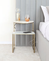 A modern two-tiered round side table with a white marble top and gold-finished metal frame, positioned next to a bed with grey padded headboard on a textured beige carpet. The upper tier supports a decorative lamp and a stemless wine glass, while the lower tier holds a smartphone with a charging cable.