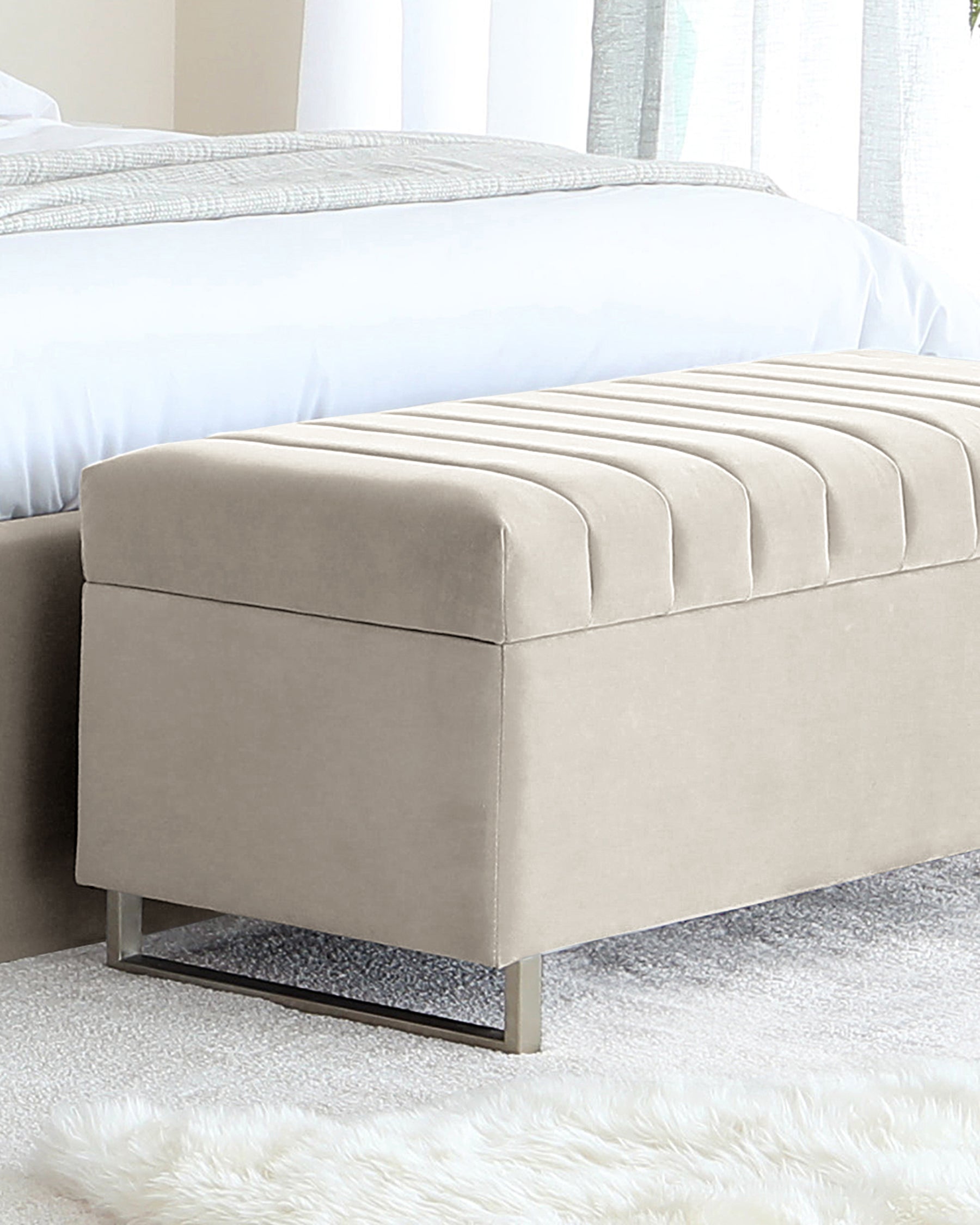 Beige upholstered bedroom bench with tufted cushion top and sleek metallic legs, positioned at the foot of a bed with a textured white comforter over a shaggy white rug.