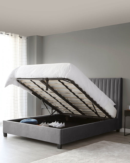 Gray upholstered storage bed with tufted headboard and gas-lift mechanism revealing under-mattress storage, set in a room with light grey walls and sheer curtains.