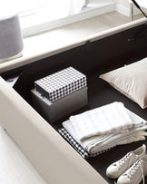 Modern minimalist style storage bed with an open bench-style footboard featuring a built-in dark upholstered seat and a storage area beneath, accented with a neutral toned throw blanket and decorative storage boxes. A sleek bedside lamp is also visible.