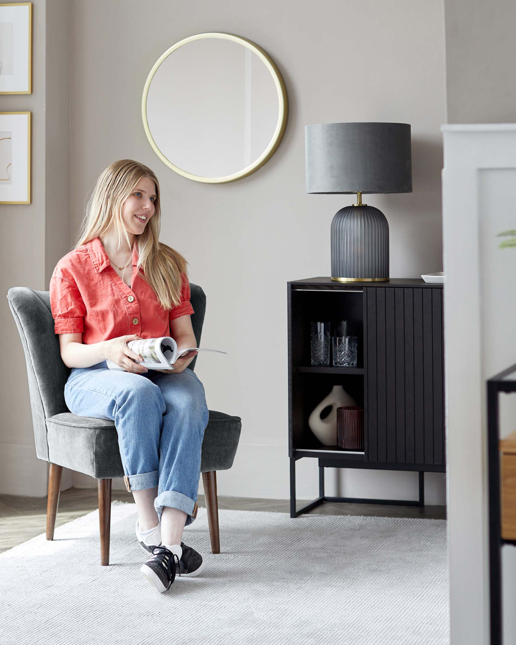 Elegant grey upholstered wingback chair with wooden legs, a sleek black sideboard cabinet with vertical grooves and brass handles, and a contemporary table lamp with a grey shade and ribbed, dark base on top of the sideboard. A circular mirror with a thin, gold frame adorns the wall.