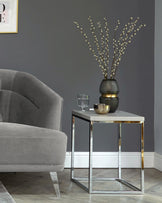 Elegant living room setup featuring a plush velvety grey armchair with a curved back and a modern side table with a sleek chrome frame and a white marble top. A decorative black and gold vase with tall pussy willow branches sits atop the side table, complemented by a simple clear water glass.