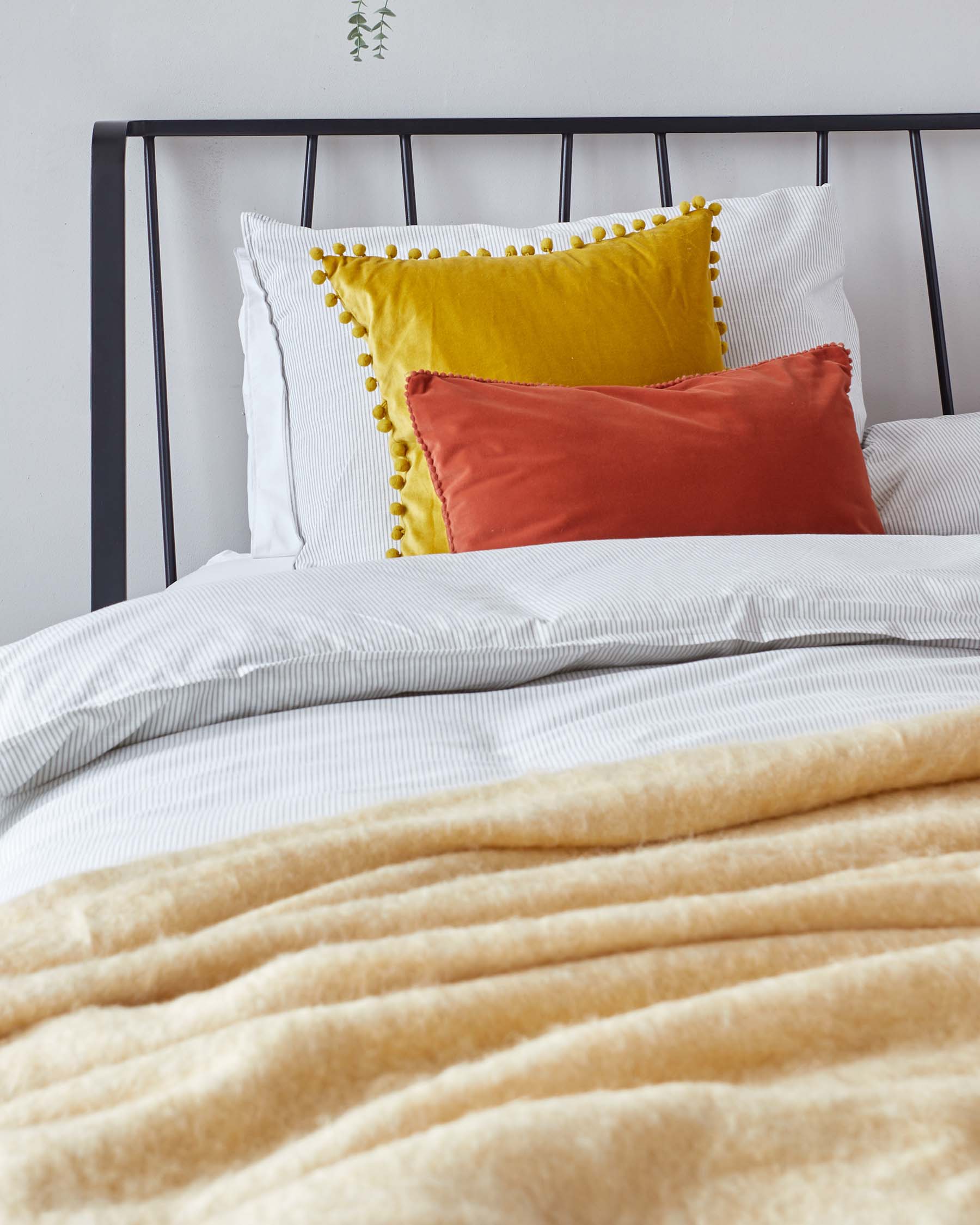 A minimalist black metal bed frame with a simple headboard design, adorned with crisp white bedding, including a textured duvet cover and pillowcases. The bed features a decorative golden-yellow pillow with pom-pom trim and a rust-coloured accent pillow in front. A plush beige throw blanket is draped across the foot of the bed, adding a layer of warmth and colour contrast.