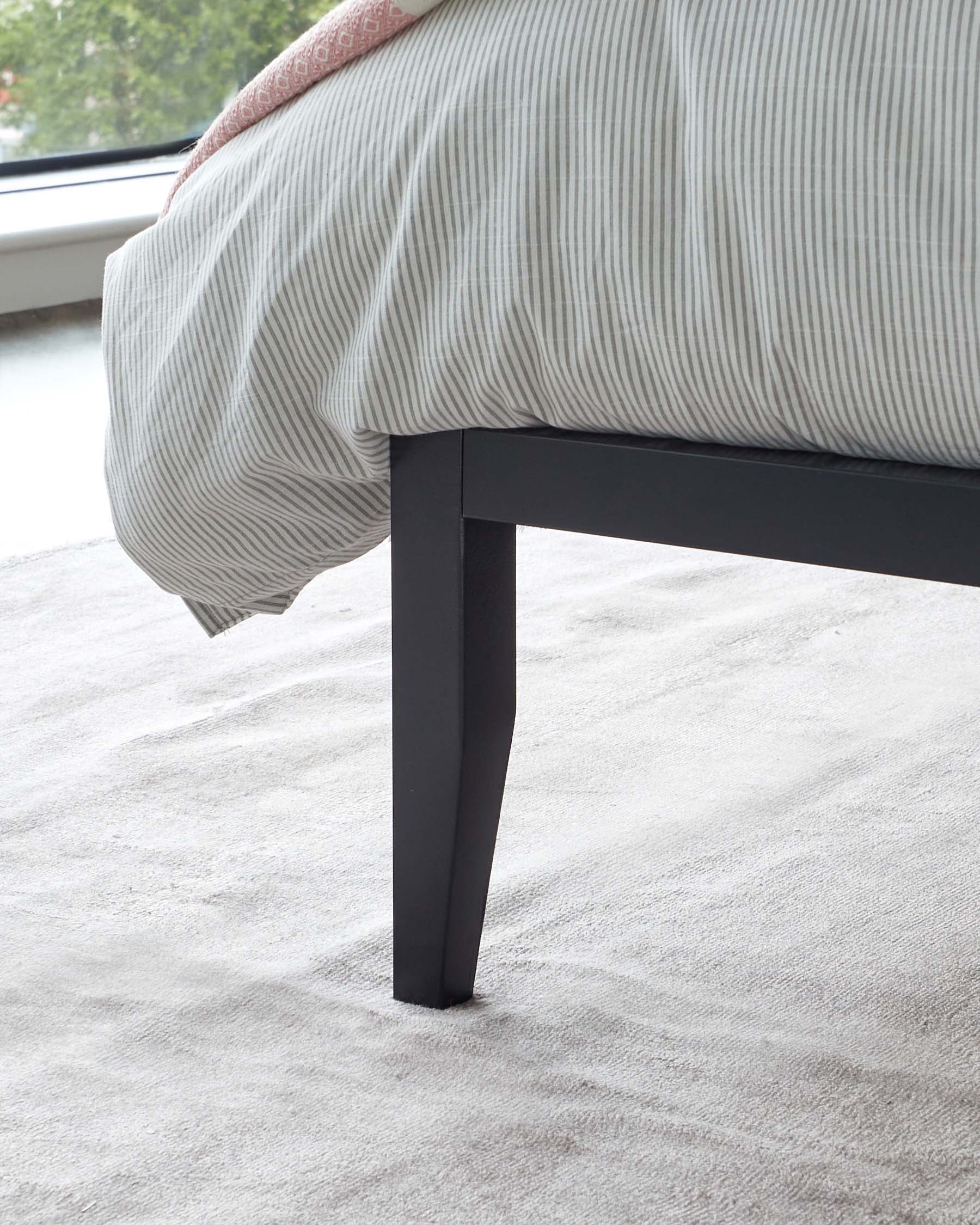 Close-up of the corner of a minimalist-style bed frame with a black, tapered leg. The bed is partially dressed with a light-coloured, striped duvet. The frame is situated on a plush, light grey carpet in a bright room with natural light.