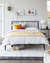 Modern black metal bed frame with layered bedding, decorative pillows, nightstand lamp, and woven basket under bed.