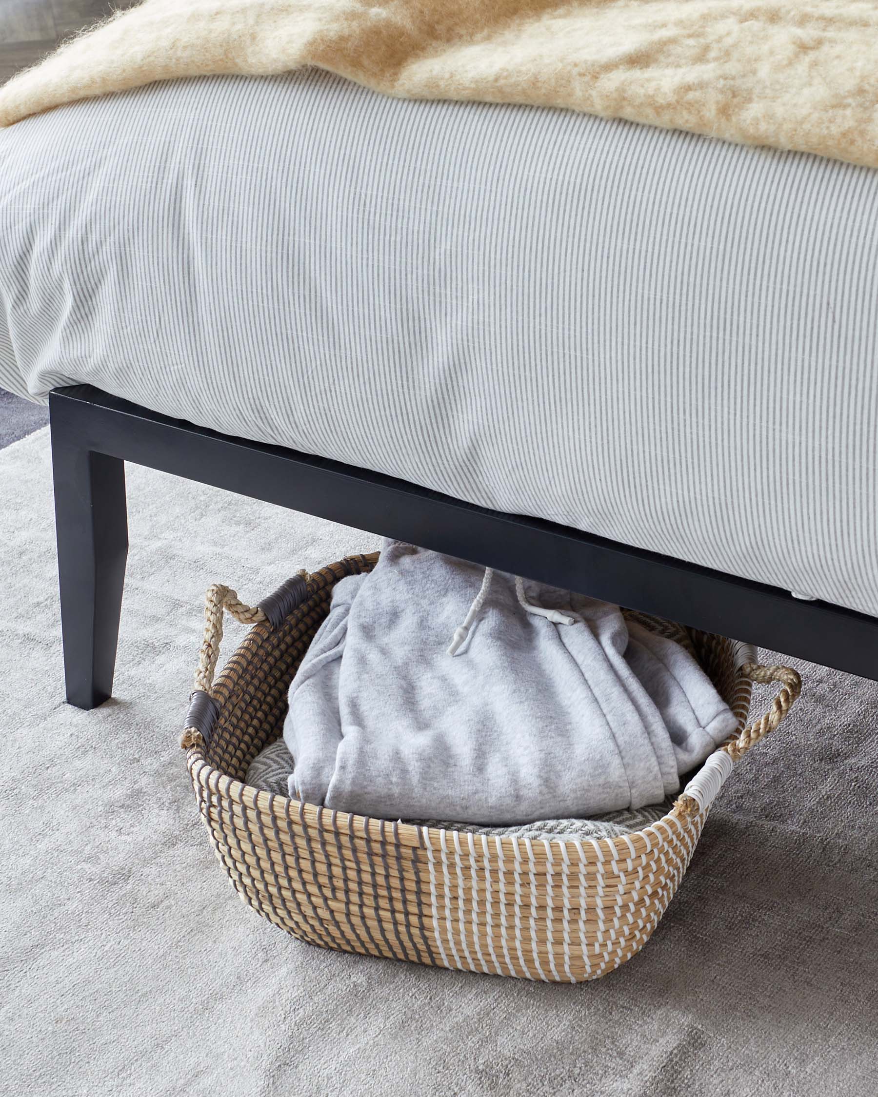 A modern minimalist black bed frame with a portion of the bed covered in a striped grey and white bedding and a tan faux fur throw. Below the bed is a woven natural fibre basket holding a folded grey sweatshirt. The setting is on a textured grey carpet.