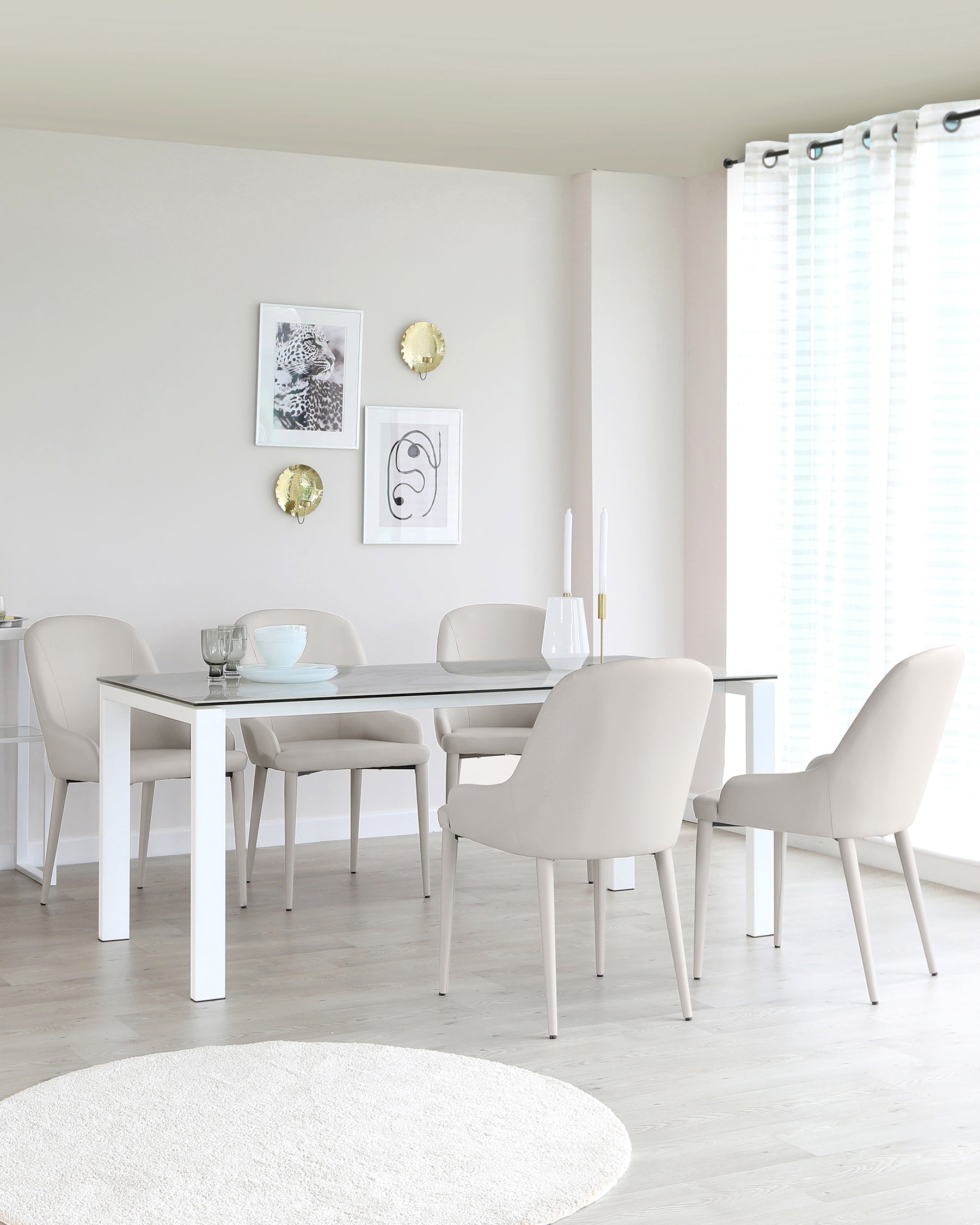 A modern dining room featuring a simple white rectangular dining table paired with four beige upholstered chairs with a minimalist design. A round, off-white area rug lies on the light wooden floor, complementing the neutral palette of the room.