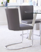 Modern dining set featuring two grey faux leather chairs with high backs and curved chrome cantilever bases, paired with a round glass-top table with a white pedestal base.