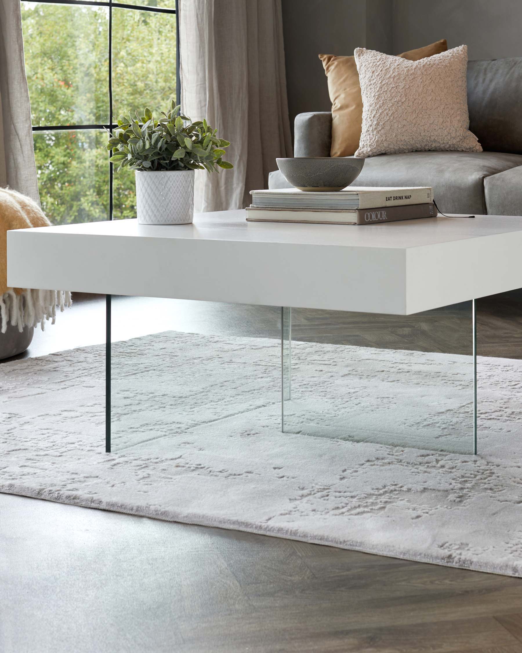 Modern minimalist white rectangular coffee table with clear glass legs on a textured grey area rug, accompanied by decorative items including a potted plant, a grey bowl, and stacked books.