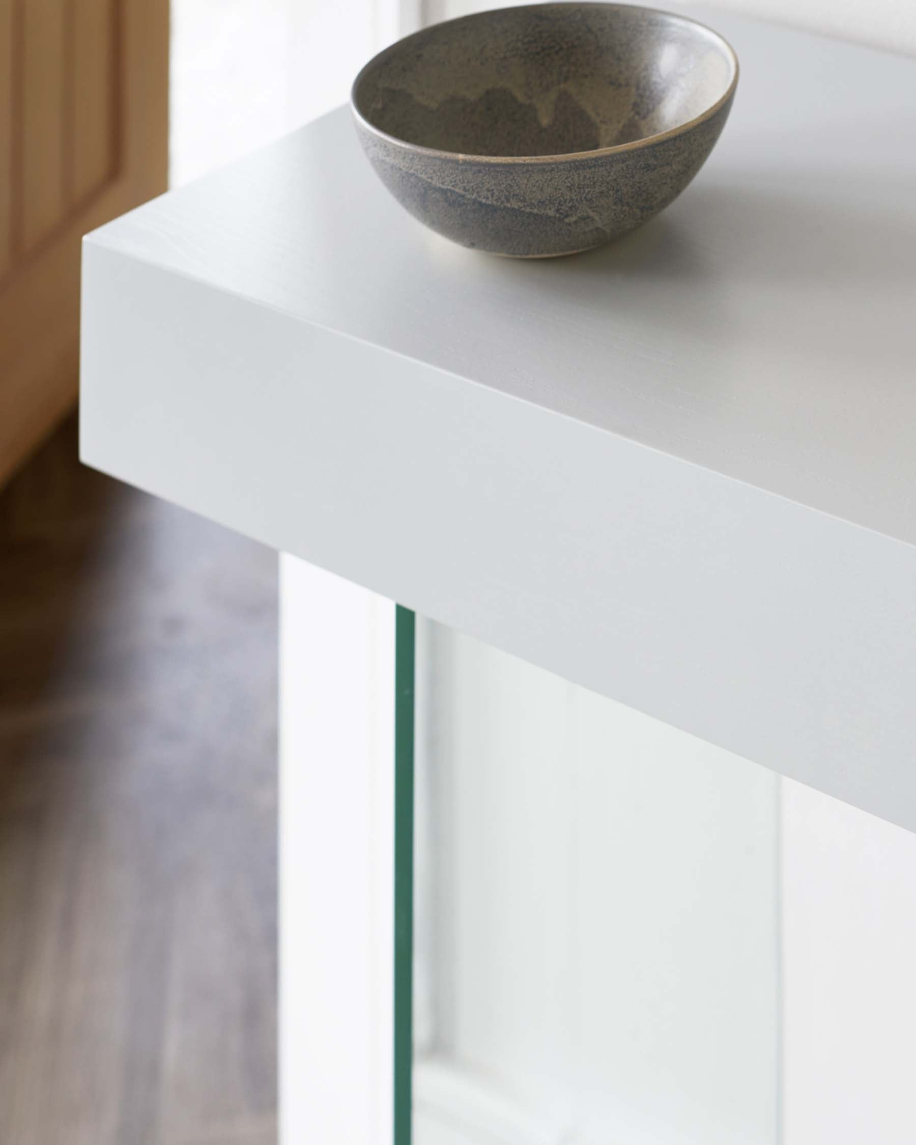 A minimalist modern white console table with clean lines, featuring a rectangular tabletop with a textured ceramic bowl placed on top. The table has a reflective surface and sleek finish, with visible clear glass side supports providing a contemporary aesthetic.