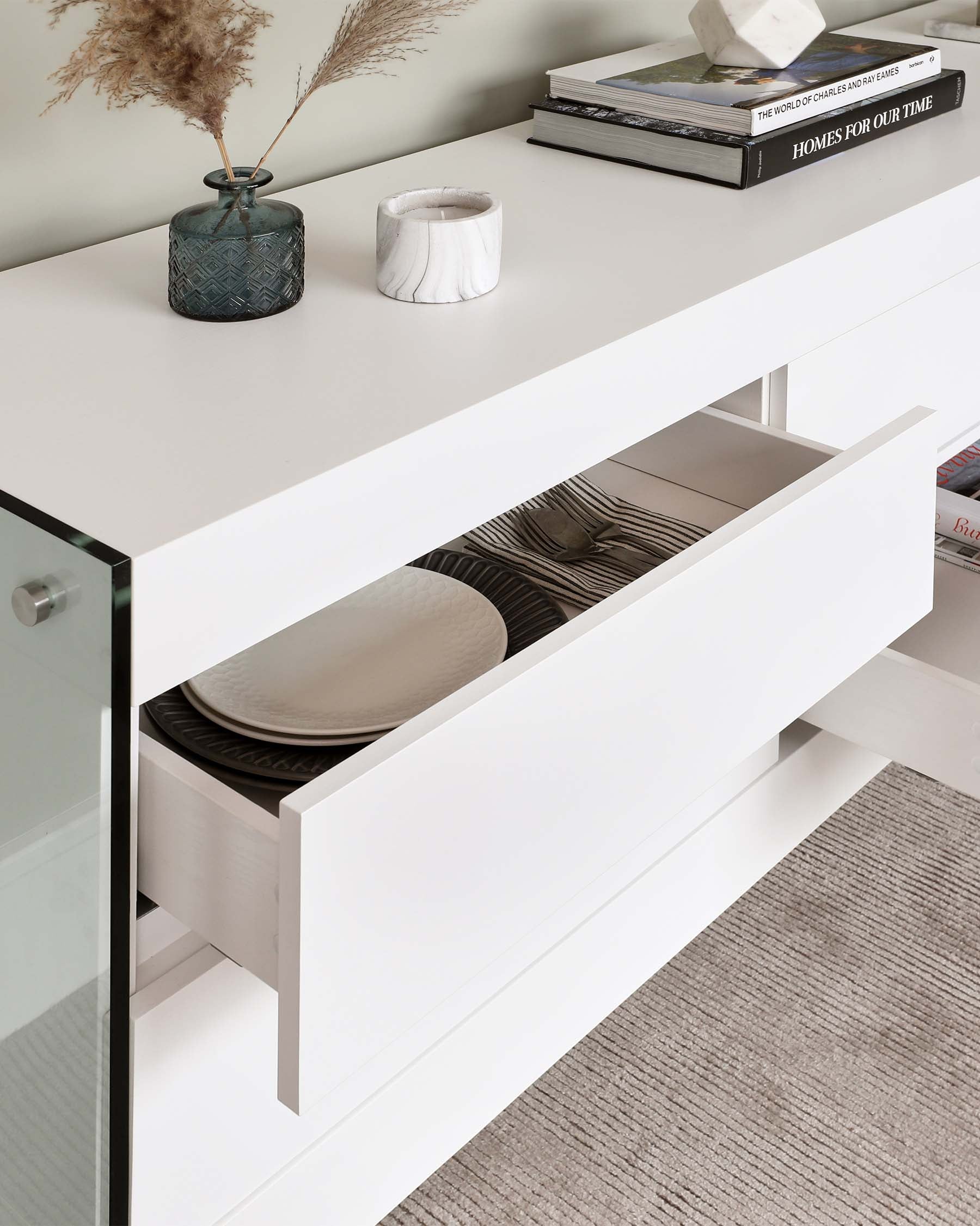 Modern minimalist white buffet with sleek lines, featuring glass side panels and a partially open drawer revealing organized dinner plates. The surface is accessorized with a textured blue vase with dried pampas grass, a white candle holder, and a stack of decorative books.