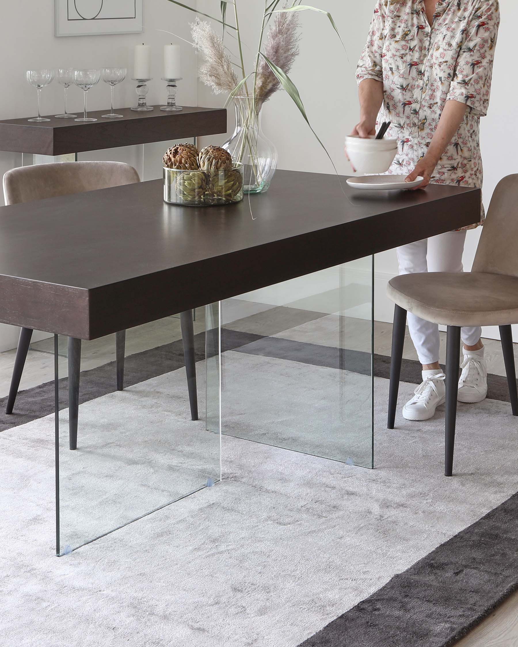 Modern rectangular dining table with a dark wood finish and clear glass legs, accompanied by velvety taupe upholstered dining chairs with black metal legs, set on a grey ombre area rug. A matching dark wood console table with decorative items is visible in the background.