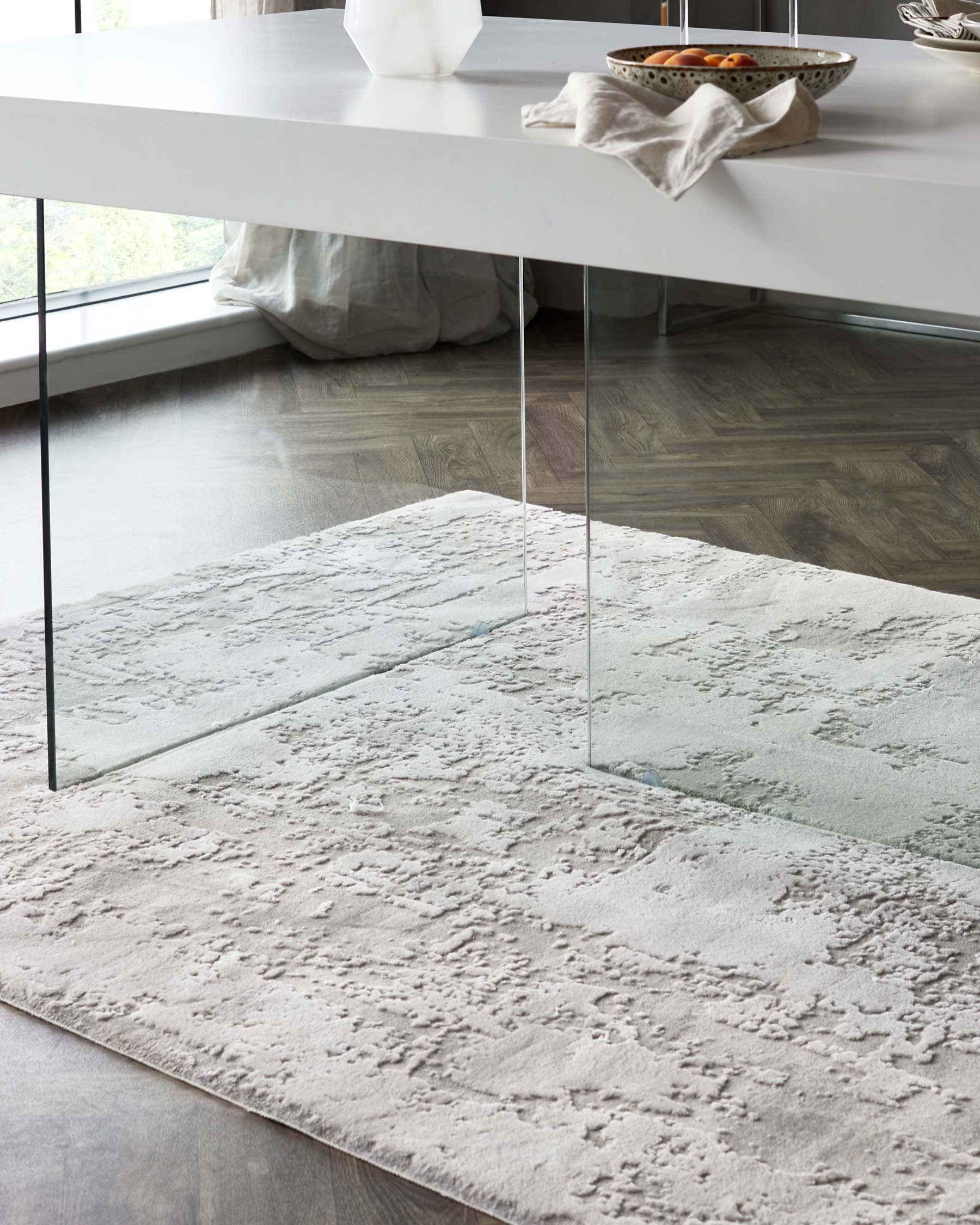 Modern minimalist dining table with a white top supported by transparent glass legs, partially set with a decorative bowl and napkin, over a textured light-coloured area rug.