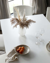 Modern minimalist white dining table with matching white chairs. The table is set with a ceramic vase containing dried pampas grass, two clear wine glasses, and a speckled ceramic bowl with fruit, accompanied by a natural linen napkin. Elegant and simple design suitable for contemporary dining spaces.