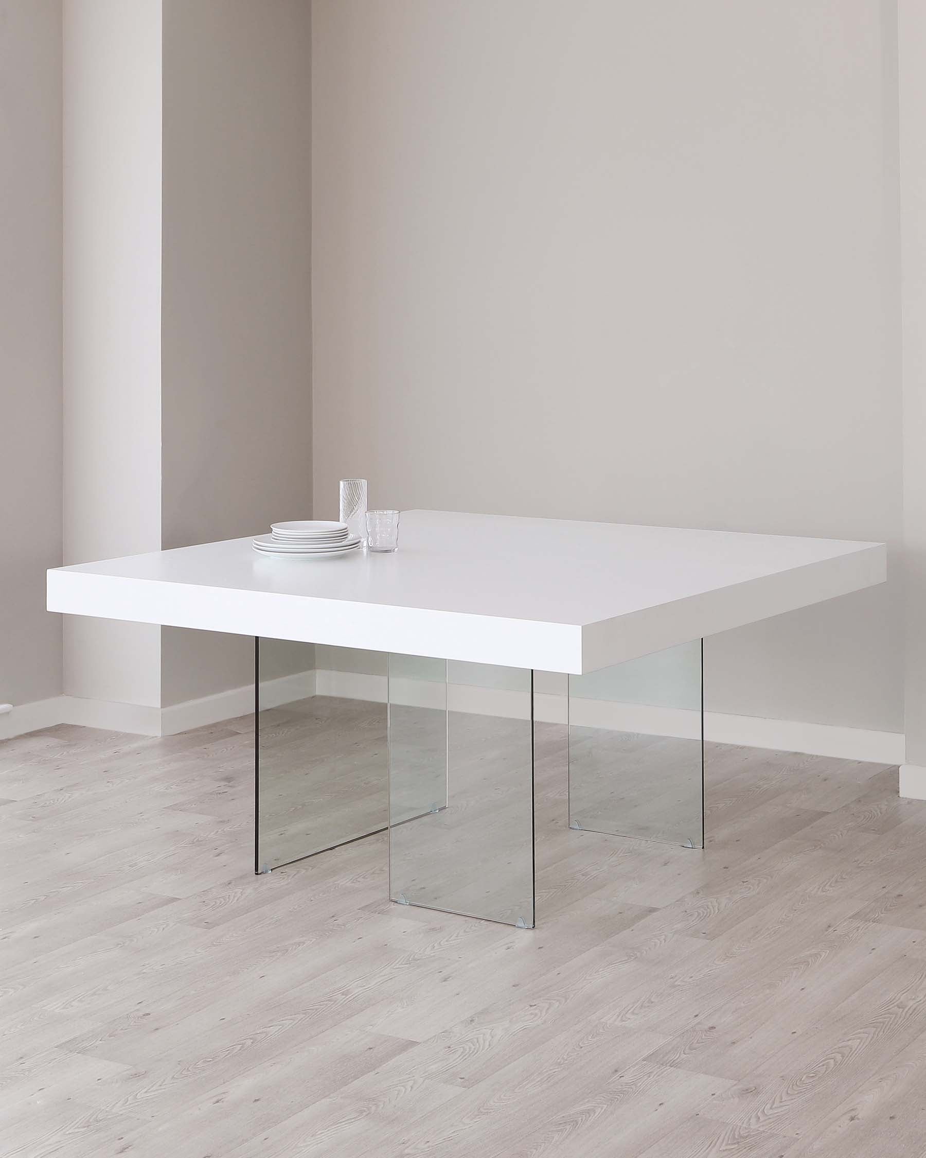 Modern minimalist white dining table with clear glass legs. Tabletop is accessorized with a simple set of white plates and transparent drinking glasses. The table stands on a light wooden floor against a plain light-coloured wall.