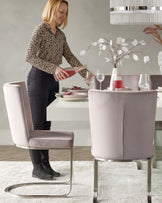 Elegant modern dining room featuring a round, white, minimalist-style table and four sleek blush pink velvet chairs with a unique curved back design and shiny chrome sled bases.