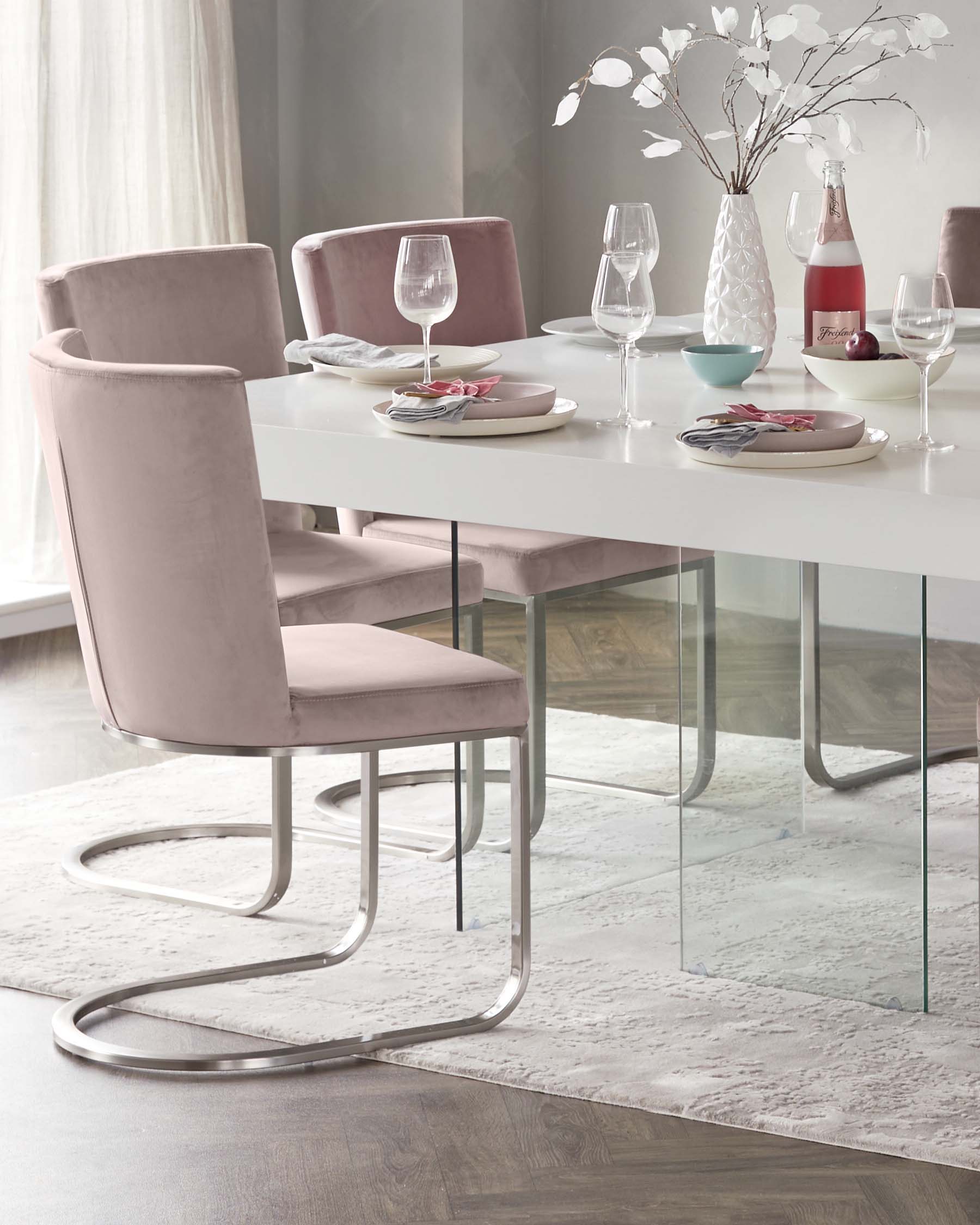 A modern dining set featuring a white rectangular table with a glossy finish and clear glass legs. Accompanied by four blush pink velvet upholstered chairs with unique curving chrome metal base, set on a textured off-white area rug.