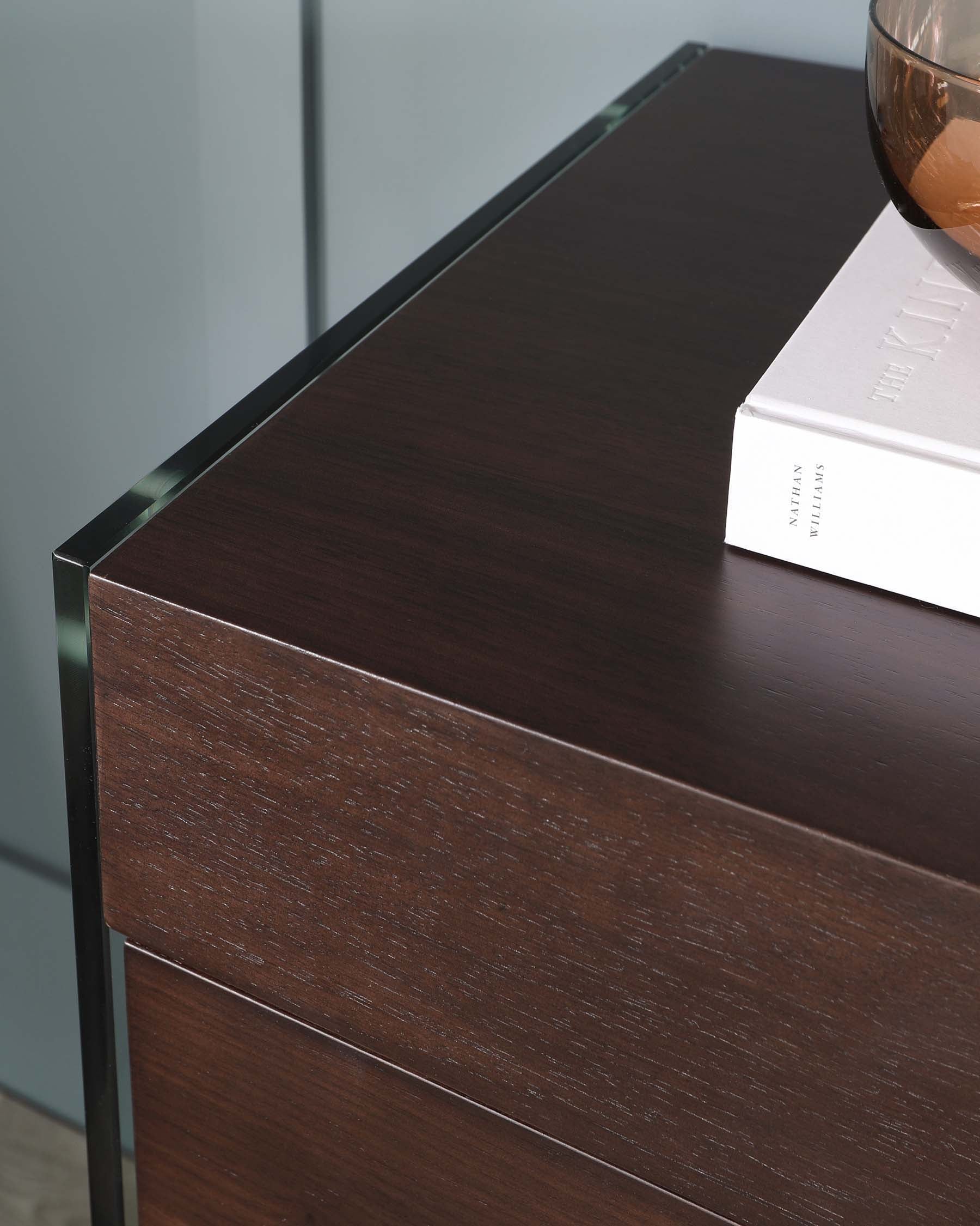 A modern dark wooden chest of drawers with a glossy finish and a glass edge detail on the top surface. There is also a hardcover book and a decorative glass bowl placed on top of the chest.