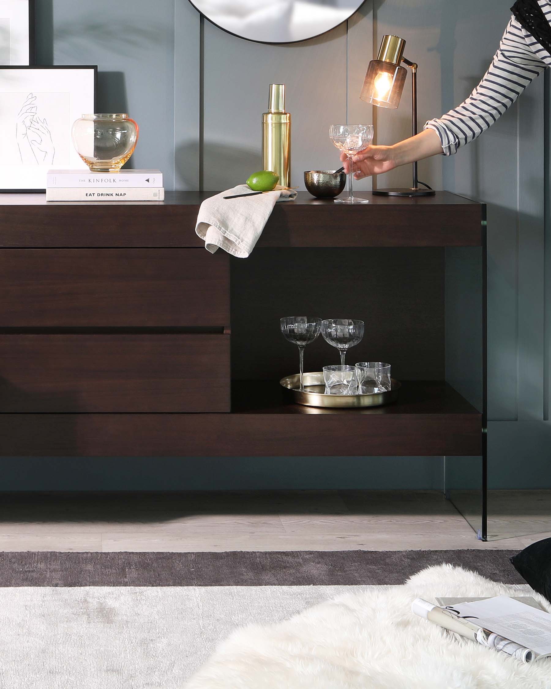 A modern, dark wood sideboard with clean lines and minimalistic hardware. The sideboard has two visible tiers of shelving, the lower featuring a round, silver tray with glassware, while the top tier is adorned with a pair of books, an artistic glass bowl, and a gold cocktail shaker, indicating sophisticated and functional styling. To the right, a contemporary lamp with a clear base and gold accents illuminates the space.