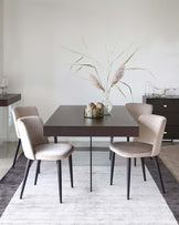 A modern dining room featuring a minimalist dark wood table and four curved-back chairs upholstered in a light beige fabric with dark wooden legs, set on a grey textured area rug. A sideboard with simple dark finish is visible in the background. The table is accessorized with a decorative centrepiece including tall grasses and pinecones in a clear glass vase.