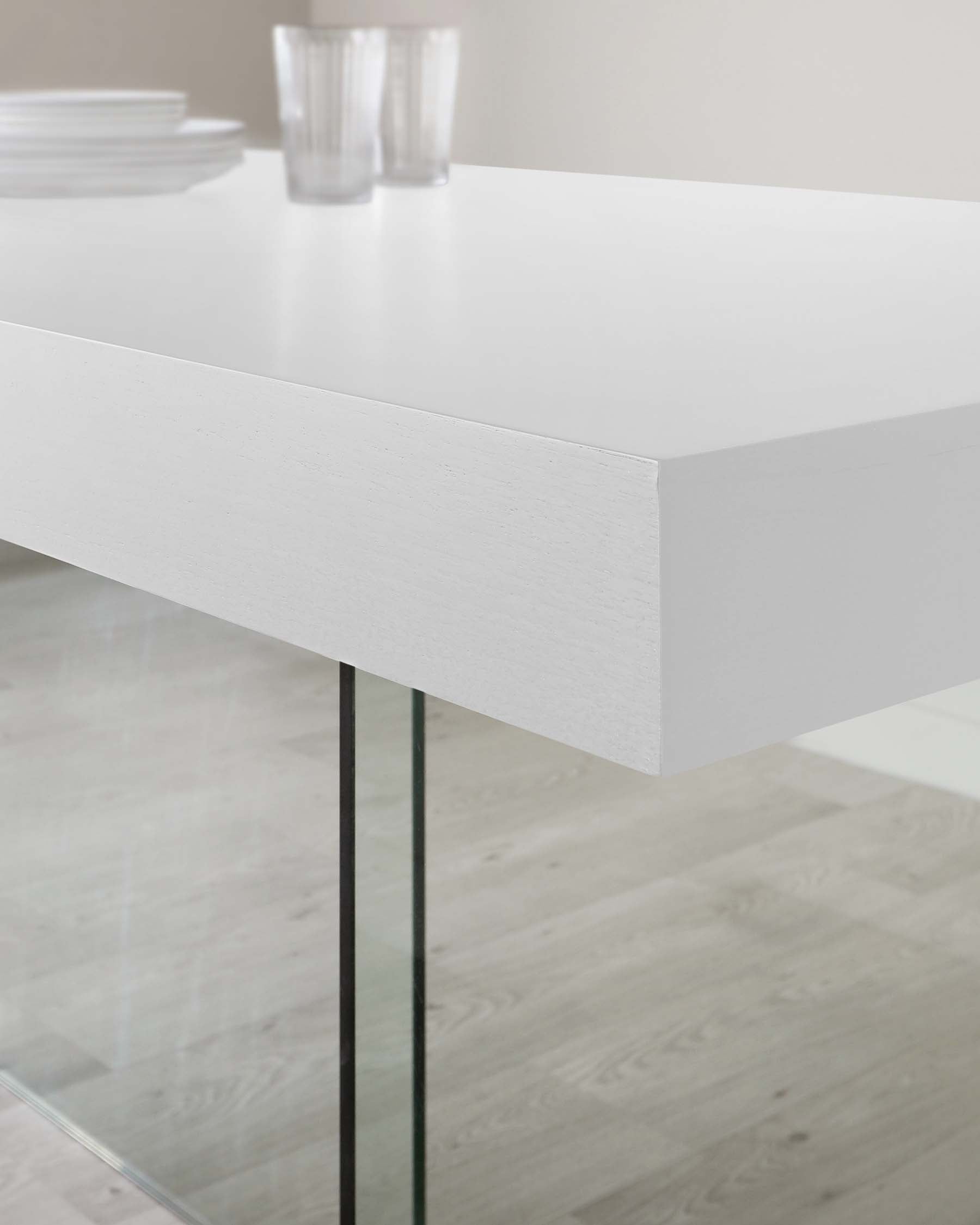 Modern minimalist white dining table with a thick, square-edged tabletop supported by a single clear glass leg, shown in a bright, neutral-toned space with subtle dishware and glassware on top.