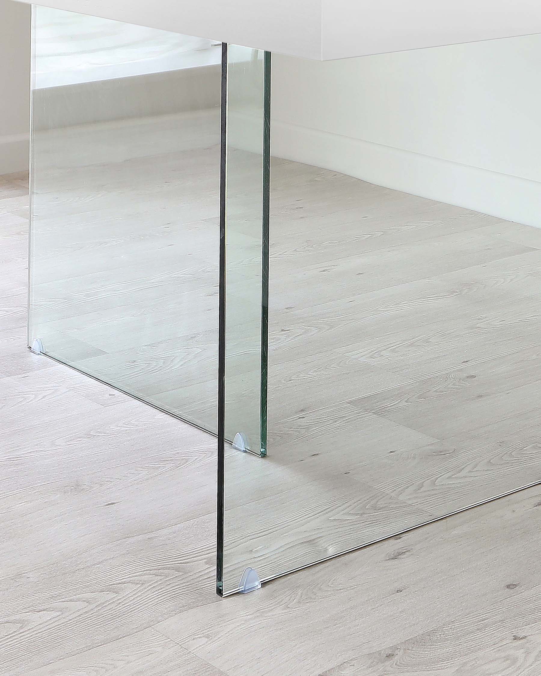 Modern minimalist glass table supported by two clear, sturdy glass panels serving as legs, set against a light wooden floor and white wall background.