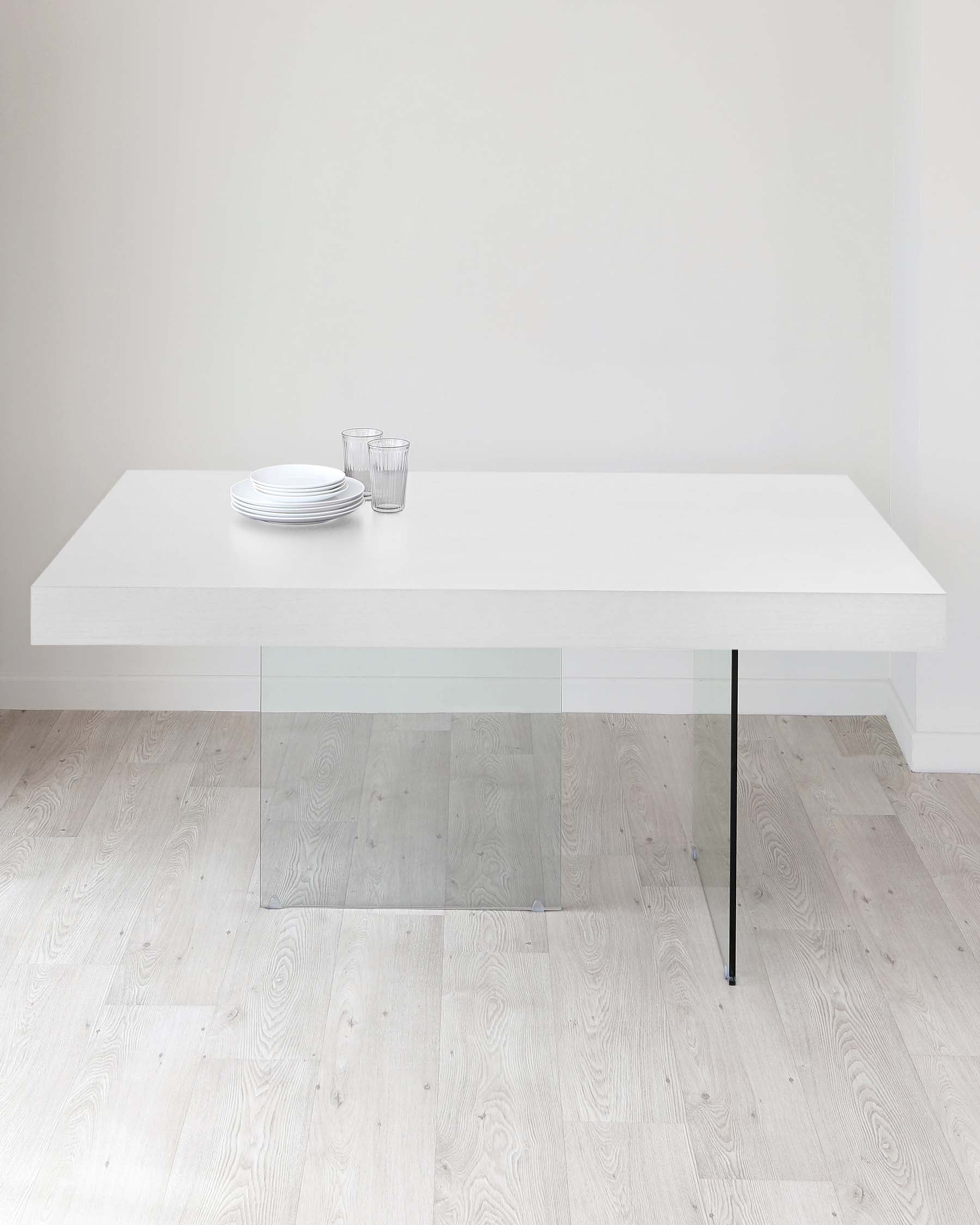 Modern minimalist white dining table with a clean, sleek design, featuring one solid leg and a transparent plexiglass support structure, on a light wooden floor. The table is set with simple white plates and clear glasses.