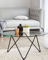 A modern grey tufted loveseat with a white rectangular pillow, paired with a round glass-top coffee table with a unique black metal base. A white marble cylindrical container holding a green plant and a small copper-coloured vase sit atop the table. The setting is complemented by a white shaggy area rug and a partial view of a striped fabric that adds a touch of pattern to the scene.