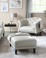 Elegant living room furniture collection featuring a cream-colored tufted leather armchair, matching ottoman, and a side table with a glass lamp and decorative items. The room has a contemporary feel with artwork and a neutral colour palette.