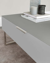 Modern minimalist grey coffee table with sleek metal handles and a glossy finish set on a neutral-toned carpet. The table is styled with stacked books and decorative candles.