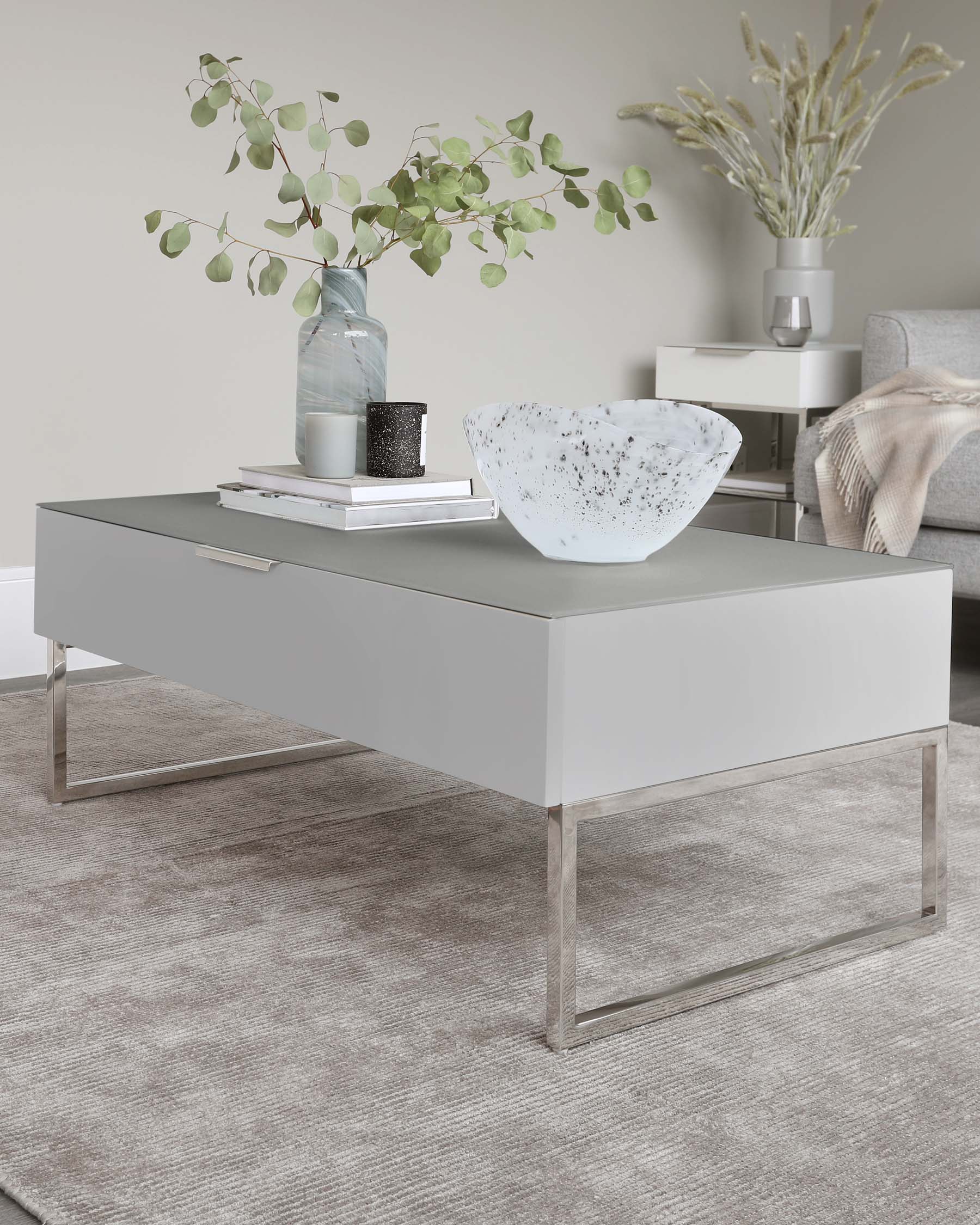 Modern minimalist living room furniture featuring a sleek rectangular grey coffee table with a glossy finish and clear acrylic legs. In the background, there is a contemporary white side table with a lower shelf, also with clear acrylic legs. Both are set against a neutral colour palette with decorative home accents on top.