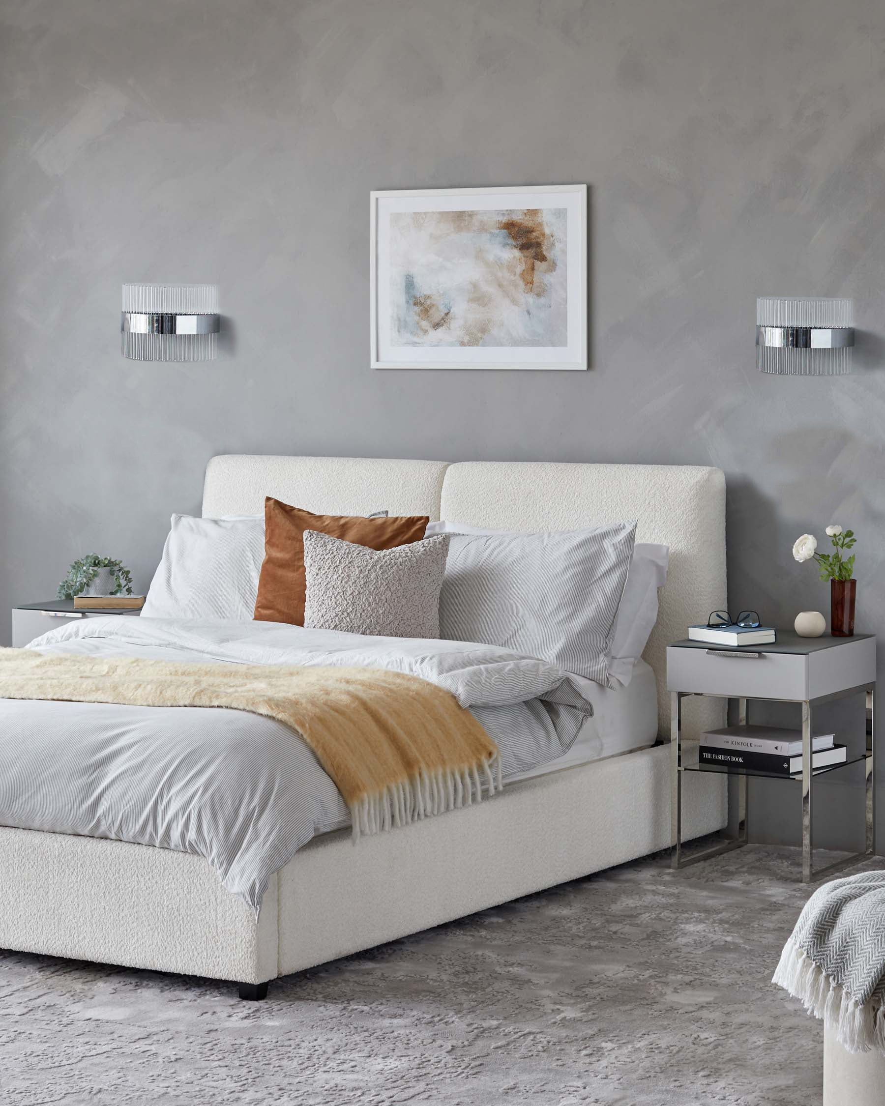 Contemporary minimalist-style bedroom featuring a king-size bed with an upholstered off-white headboard and a matching bed frame. The bed is dressed with white bedding, accented with a soft beige throw and decorative pillows in earth tones. Beside the bed is a modern grey bedside table with clean lines and a simple metal frame, hosting a small selection of decorative items and books on its open lower shelf.