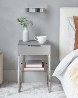 Modern minimalist bedside table with a sleek grey finish and chrome frame, featuring a single drawer and a lower shelf with two stacked books. The table is adorned with a textured ceramic vase containing eucalyptus sprigs and a white ribbed cup and saucer set.