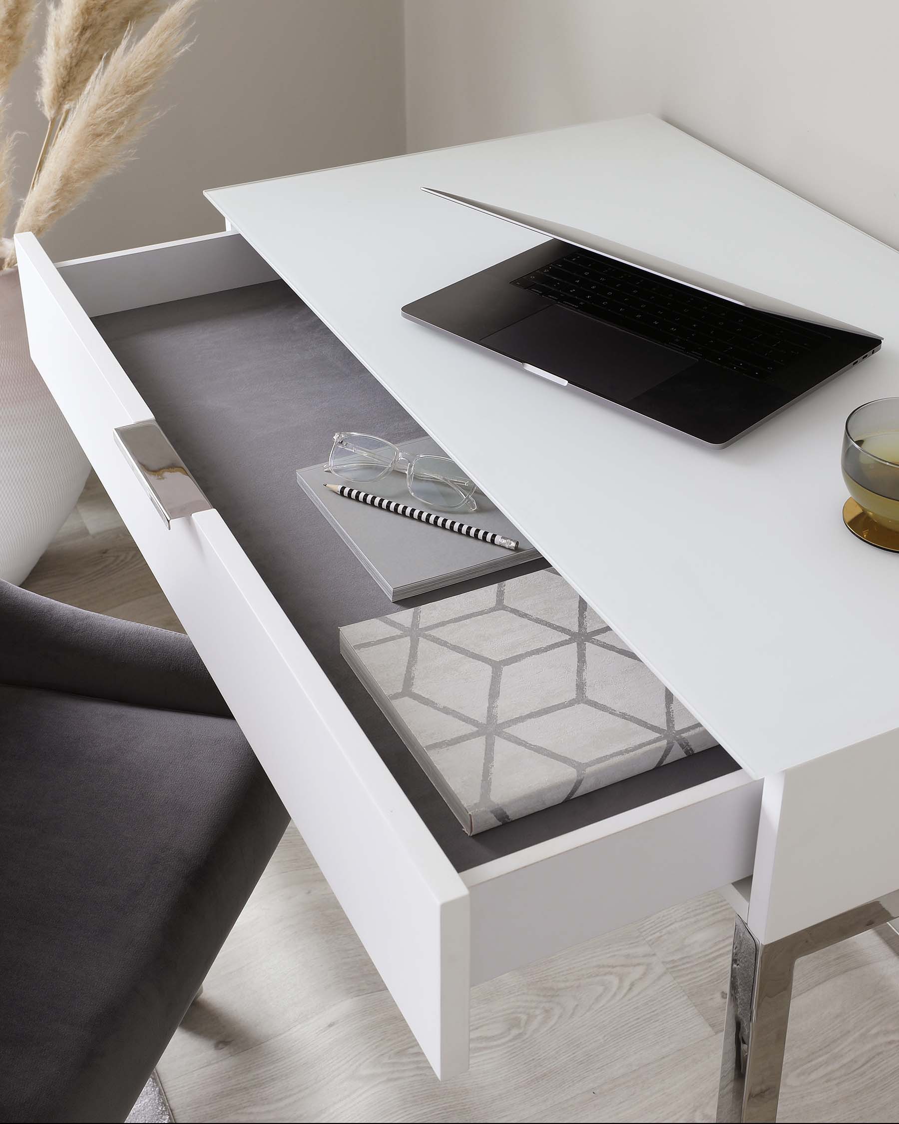 Modern white desk with an open drawer showcasing organized storage space, paired with a grey upholstered chair.