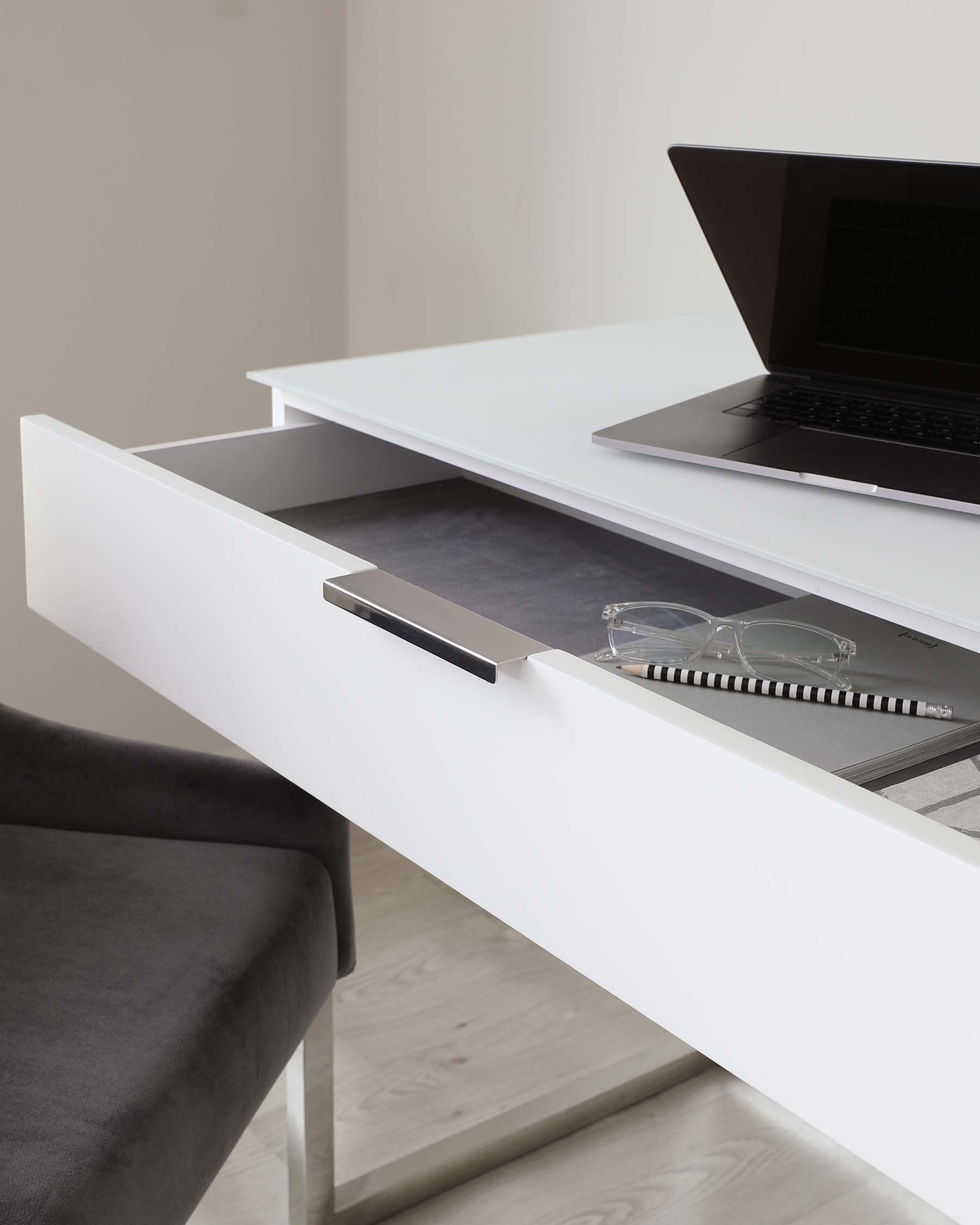 Modern minimalist white desk with an open drawer showing storage space, paired with a dark grey upholstered chair with metal legs, and a laptop placed on the desk surface.