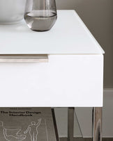 Modern minimalist white bedside table with a discrete drawer handle, standing on sleek metallic legs, partially displaying a design book underneath.