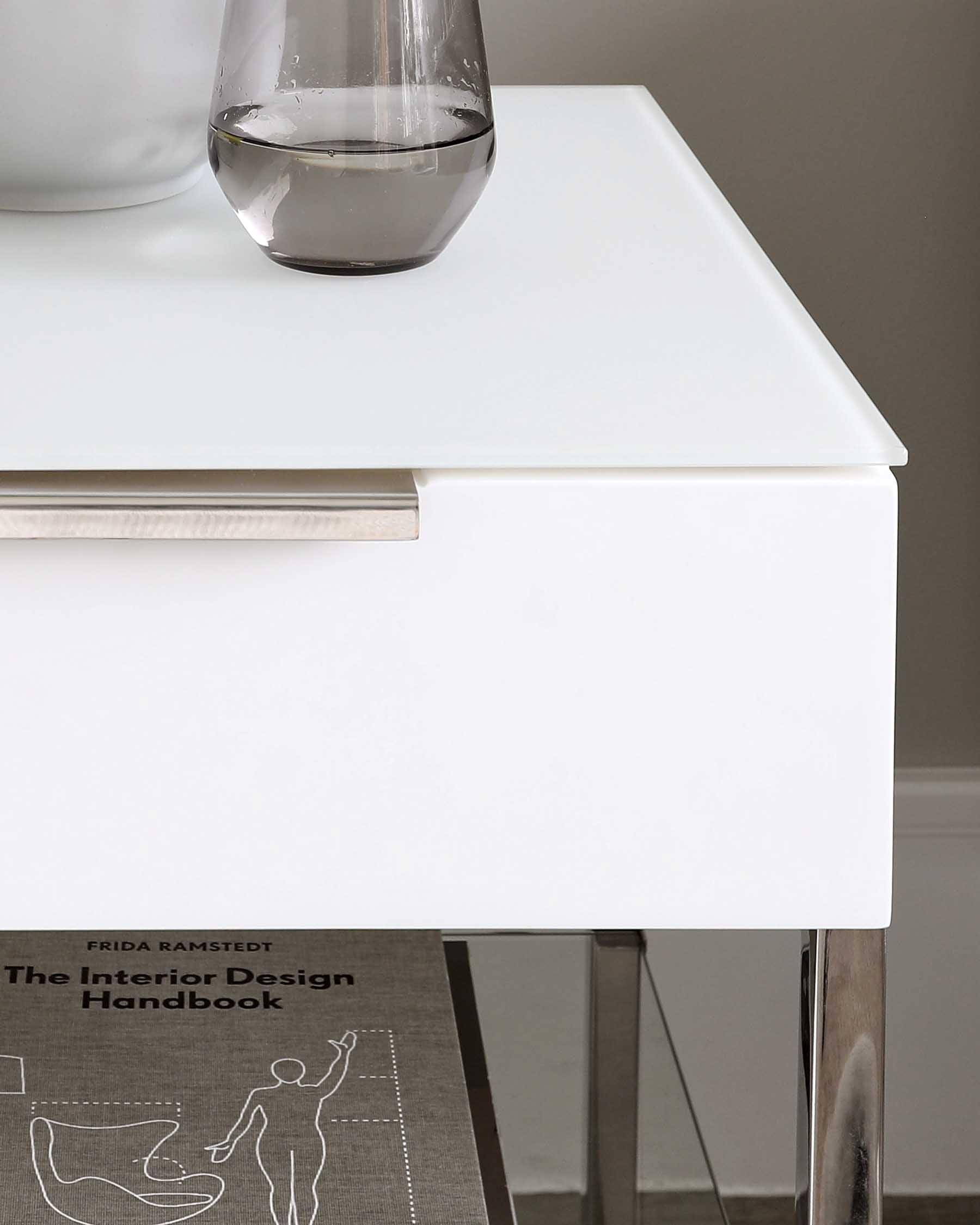 Modern minimalist white bedside table with a discrete drawer handle, standing on sleek metallic legs, partially displaying a design book underneath.