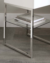 Modern minimalist glass side table with a polished stainless steel frame, featuring two levels for display and storage, complemented by a stack of design books on the upper shelf.