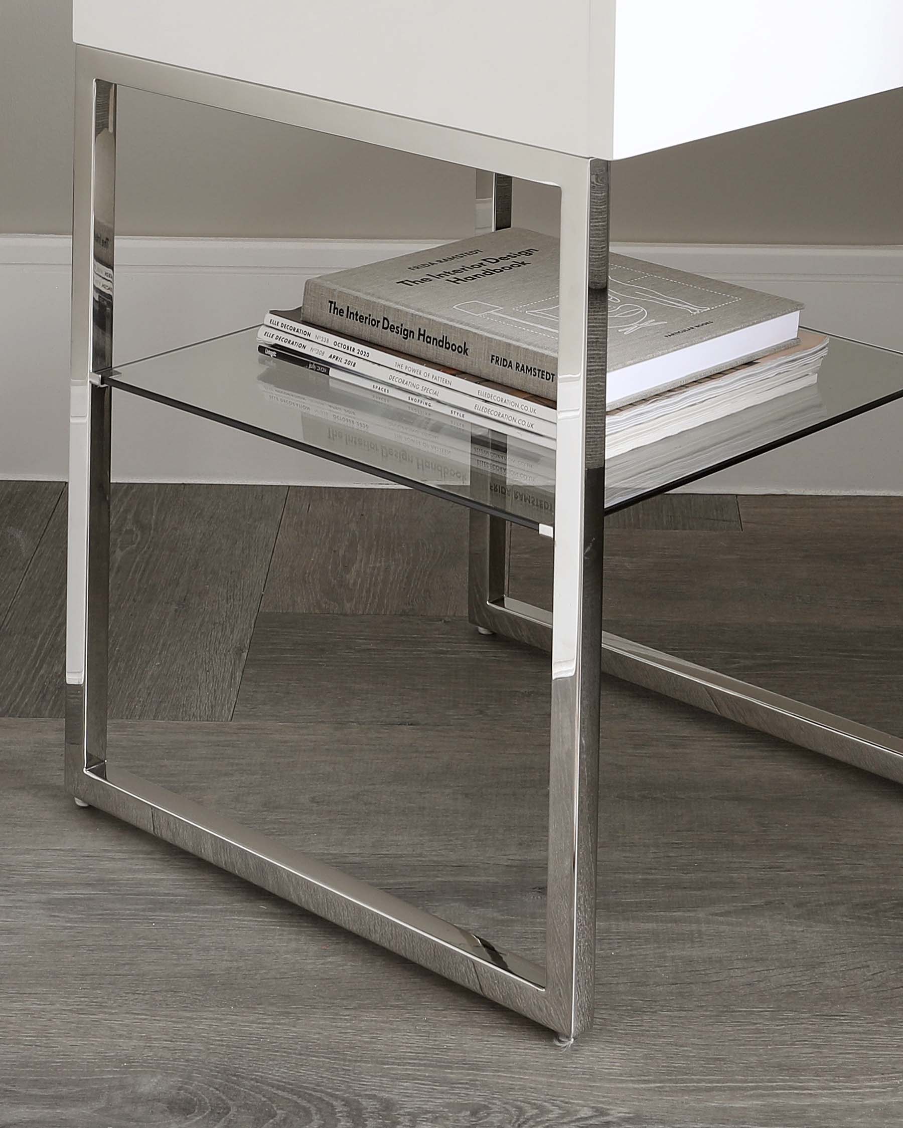 Modern minimalist glass side table with a polished stainless steel frame, featuring two levels for display and storage, complemented by a stack of design books on the upper shelf.