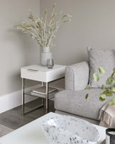 A modern white side table with a sleek drawer and stainless steel legs, paired with a textured light grey upholstered armchair. The scene is accented by a matte grey vase with dried plants on the table, adding a touch of nature to the sophisticated, minimalist decor.