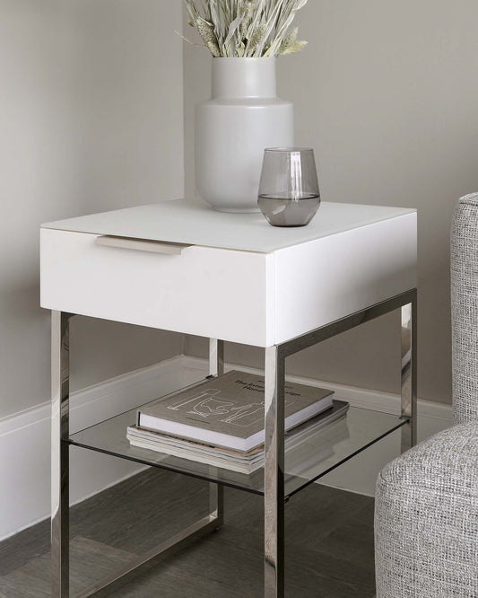 Modern bedside table with a white, square top drawer, sleek silver handle, and stainless steel frame. Features a lower glass shelf suitable for books or decorative items. Positioned next to an upholstered chair, with decorative vase and glass atop the table.