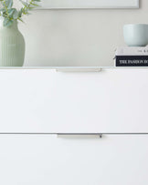 Minimalist white modern cabinet with sleek metal handles against a light grey wall, adorned with a pale green vase containing eucalyptus branches, a decorative pale blue bowl, and a stack of books, one titled "The Fashion Book".