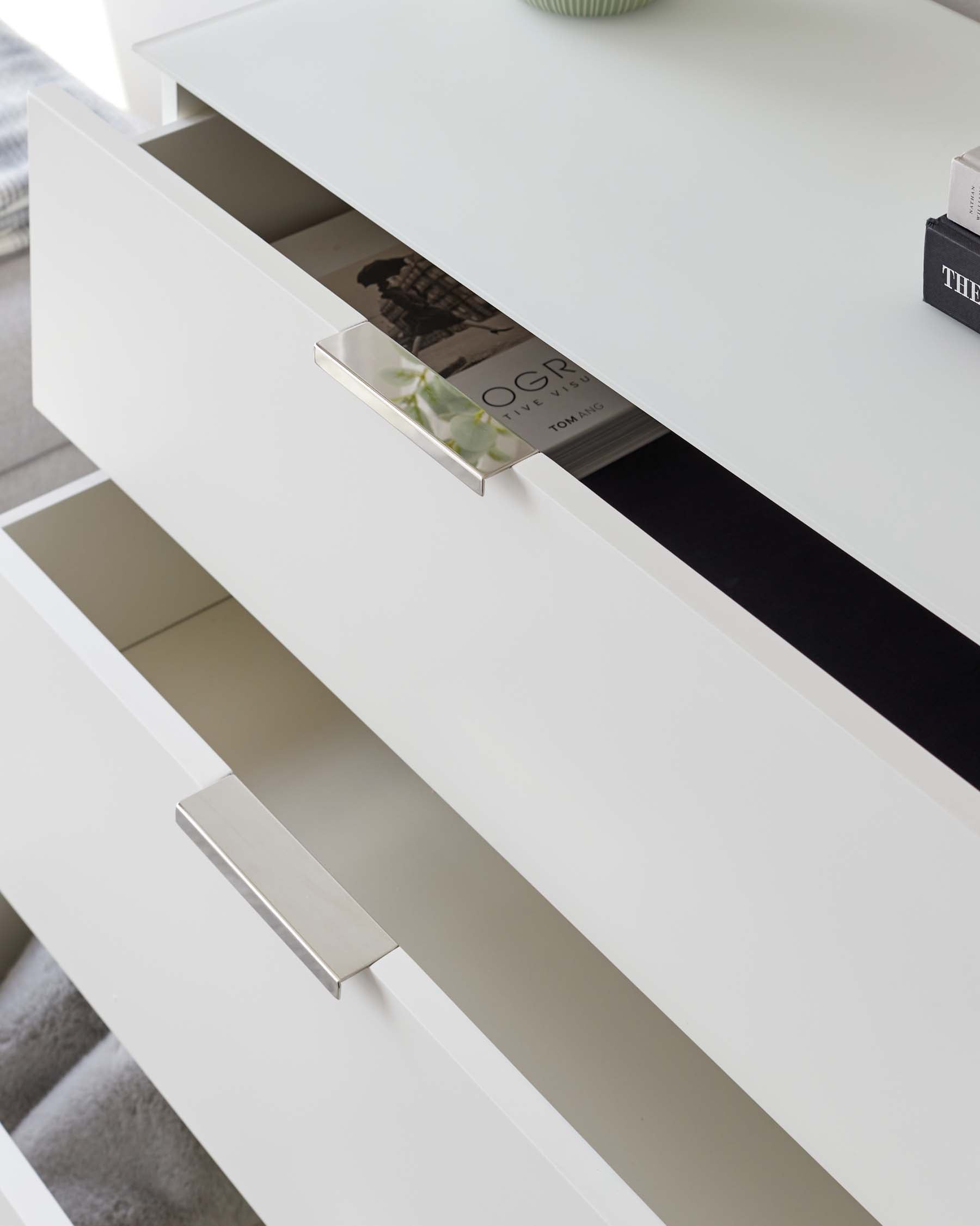 A modern white high-gloss dresser with sleek, silver-toned linear handles on the drawers. The top drawer is partially open, revealing a magazine, and the design includes a darker accent strip running horizontally along the upper edge of the drawer face.