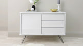 White modern minimalist sideboard cabinet with two doors and three drawers on angled metal legs in a light, neutral interior setting. A lamp, vase with greenery, stack of books, and decorative bowl adorn the top.