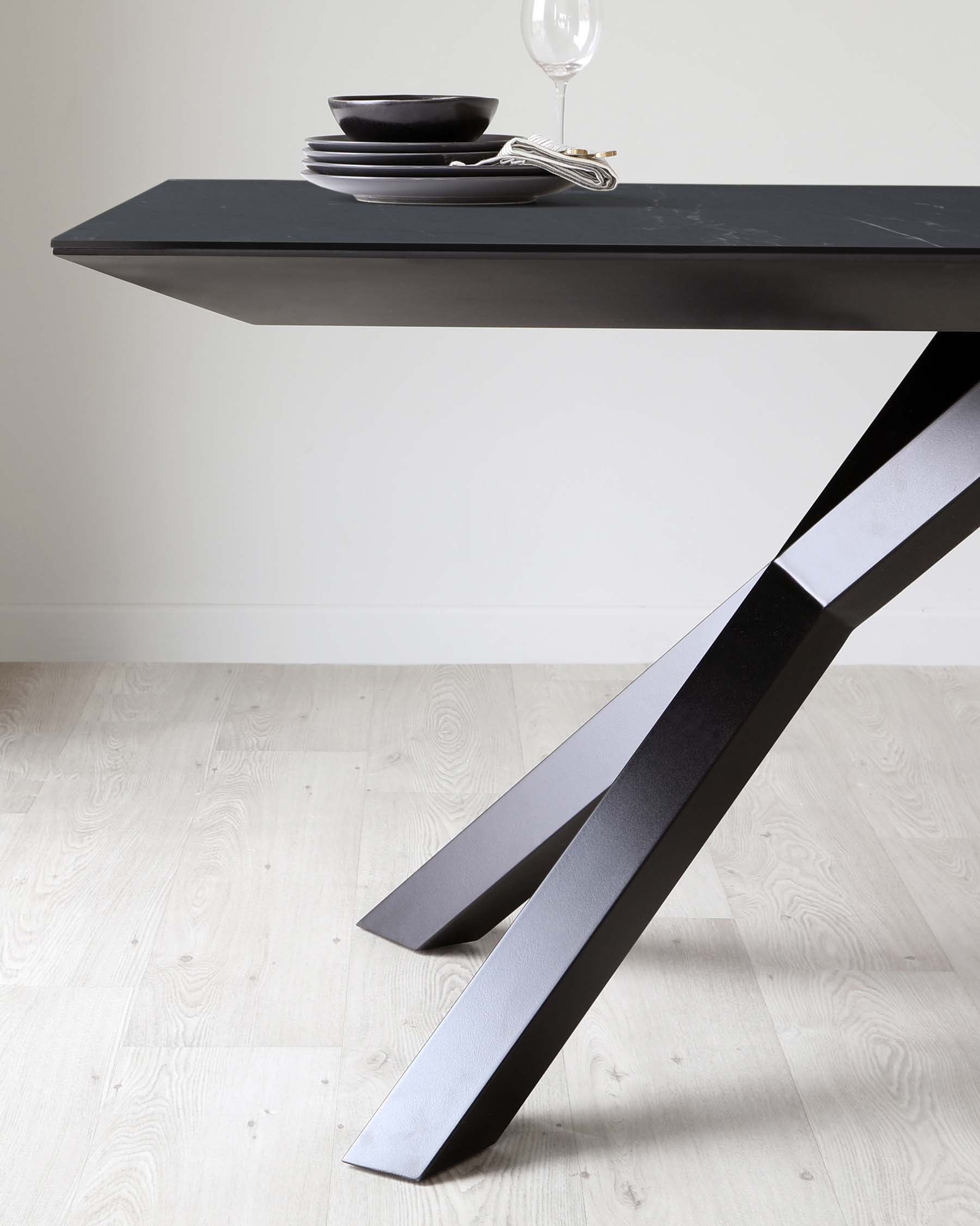 A modern minimalist black dining table with a sleek, matte finish. The table exhibits a distinctive design with wide, angled legs that taper towards the floor, providing a stable and stylish base. The tabletop is adorned with a simple table setting featuring a stack of dark bowls, a folded napkin, and a clear wine glass, emphasizing the table's elegant surface. The backdrop of a neutral wall and light herringer wooden flooring complements the contemporary aesthetic of the furniture piece.