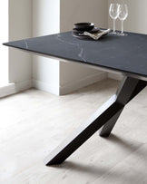 Modern minimalist dining table with a dark marble tabletop and an angular black metal base placed on a light wooden floor. The table is set with a stack of black matte plates, a folded cloth napkin, and two clear wine glasses. The room has light walls and a white skirting board.