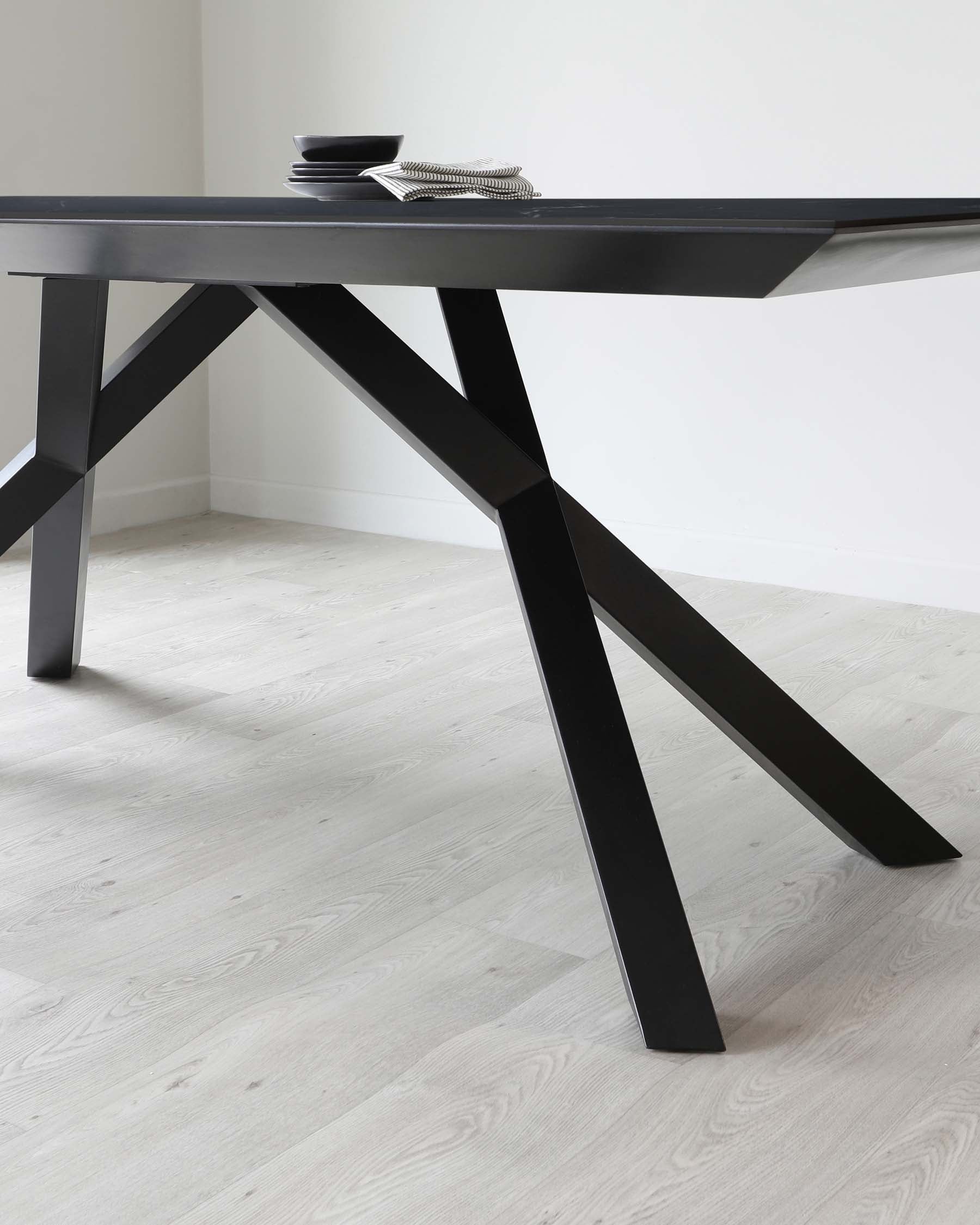 A sleek, modern black dining table with a minimalist design featuring angular legs and a thin tabletop. The surface displays a set of monochrome dishes and a silver decorative item, emphasizing the table's elegant simplicity. The table stands on a light wooden floor against a white wall, highlighting its stark geometric form and contemporary aesthetic.