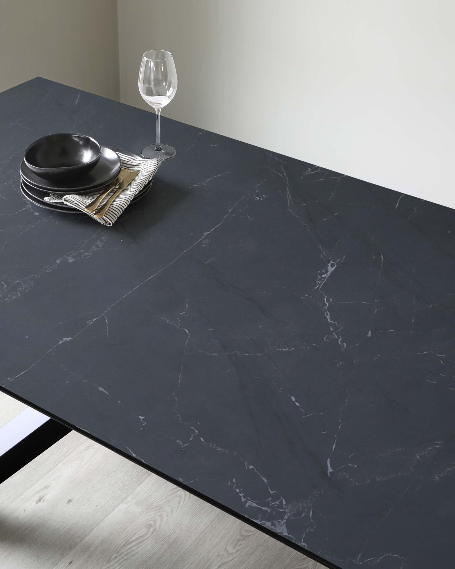 A modern minimalist dining table with a dark, marbled tabletop surface, resting on what appears to be a sleek black frame. The tabletop's expansive, unadorned surface showcases the elegant veining, giving an air of luxury and simplicity.