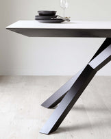 Modern minimalist white tabletop with distinctive angled matte black steel legs. On the tabletop, there's a set of matte black plates and a wine glass suggesting an elegant dining arrangement.